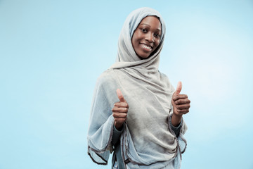 The beautiful young black african muslim girl wearing gray hijab at blue studio. She standing with...