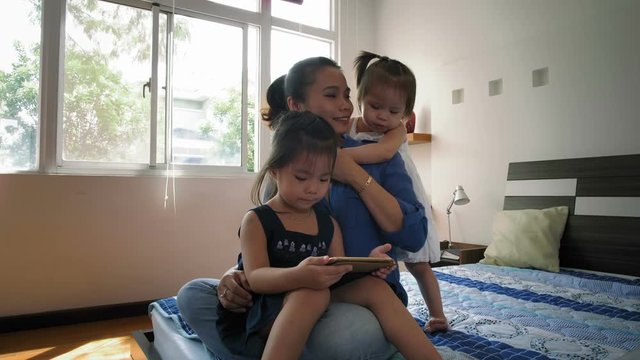 Lockdown of Asian woman sitting on bed with her daughters, hugging them, while eldest girl using tablet