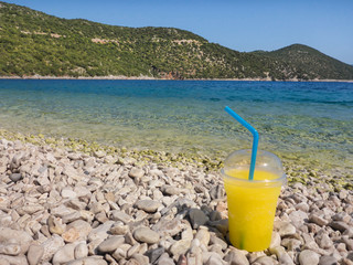 A glass of refreshing cold summer citrus lemon drink stands on white stones at pebble beach on the background of turquoise sea Antisamos beach, Kefalonia island, Greece.