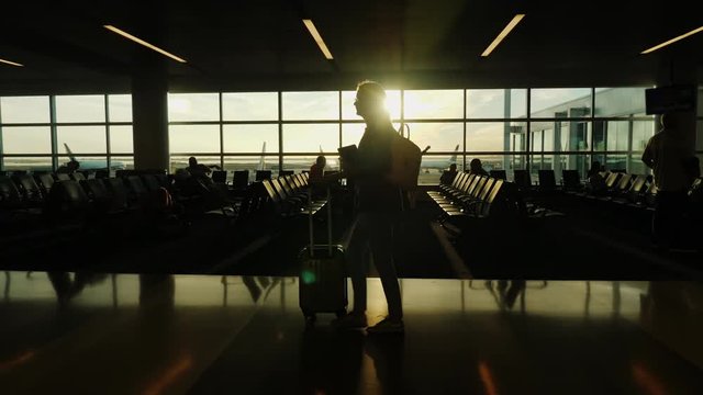 Silhouette of a woman in a suit. He goes with his luggage at the airport terminal. The setting sun shines in a large window