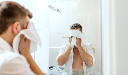 Handsome businessman with unbuttoned shirt wiping his face with towel while standing inn bathroom in front of mirror. Morning routine concept.