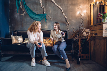 Fototapeta na wymiar Two friends or sisters enthusiastically discuss something sitting on the sofa in a cozy festive atmosphere over a glass of wine.