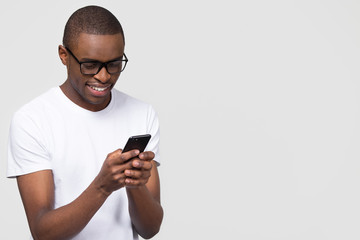 Smiling black young man in glasses holding phone looking at smartphone isolated on white grey...