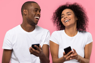 Happy young african american couple laughing using smartphones apps isolated on pink studio background, cheerful black friends users holding mobile phones gadgets having fun with cellphones together