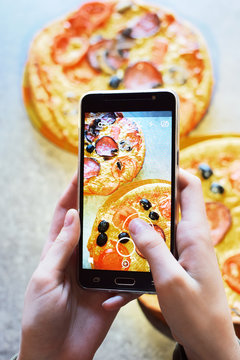 Teenage girl's hands with smartphone takes picture of fresh just baked hot selfmade pizza with salami, tomato, cheese, olives and mushrooms on grey grunge table background.