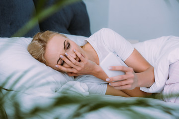 stressed woman lying in bed, covering face with hand and using smartphone