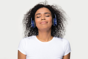 Happy calm african teen girl wearing blue headphones listening to favorite modern music isolated on white grey blank studio background, smiling black woman enjoy good sound chilling with eyes closed