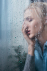 Fototapeta premium selective focus of sad woman covering face with hand through window with raindrops and copy space