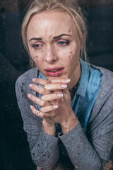 upset adult woman with folded hands crying at home through window with raindrops