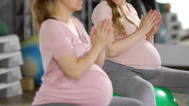 Tilt up medium shot of two fit pregnant women practicing yoga hand positions while sitting on stability balls