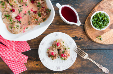 Swedish meatballs in a red cowberry sauce with green onion. White casserole and plate on wooden rustic table, top view