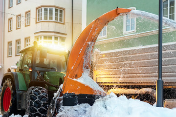Big tractor with chains on wheel blowing snow from city street into dump truck body. Cleaning...