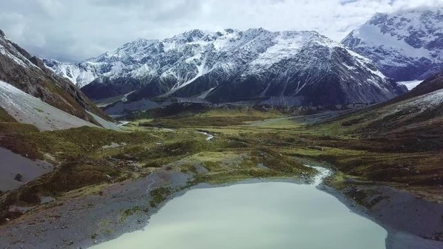 Aerial shot of shallow stream flowing into glacial lake in valley at the foot of snow-capped mountain range
