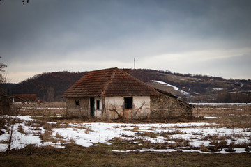 Old small abandoned house on meadow.Winter rural scene at sunset with small bits of snow.