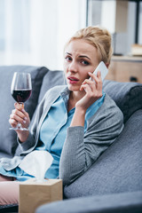 woman sitting on couch, looking at camera, holding glass of red wine and talking on smartphone at home