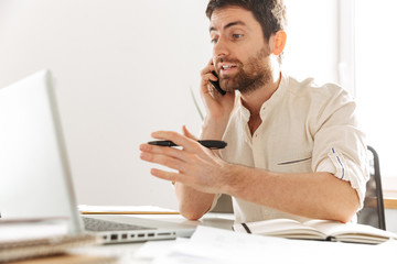Image of confident businessman 30s wearing white shirt talking on mobile phone, while sitting at table in modern office
