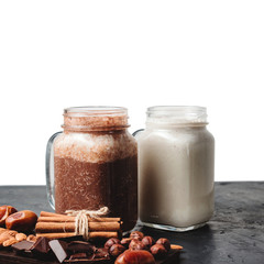 Banana and chocolate smoothie in two jars, protein milkshake, nuts, chocolate cubes, dates, cinnamon sticks on a dark table isolated on white background