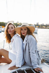 Young beautiful woman in white shirt and pretty lady in stripped jacket and straw hat thoughtfully looking in camera together on yacht with harbor view on background