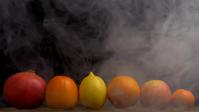 Tropical juicy fruits pomegranate, orange, tangerine, lemon and persimmon on a black background, from which blows freshness and evaporation, smoke, 4K