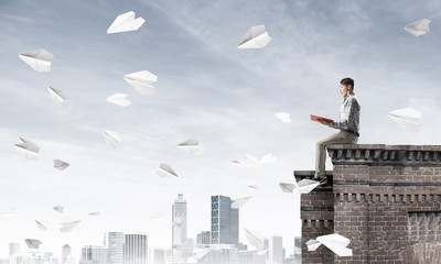 Handsome student guy on roof edge reading book and paper planes fly around