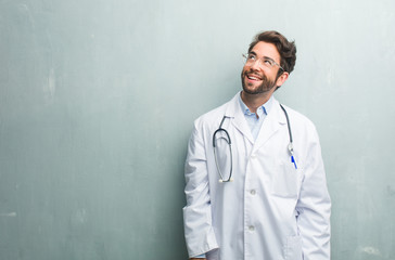 Young friendly doctor man against a grunge wall with a copy space looking up, thinking of something fun and having an idea, concept of imagination, happy and excited