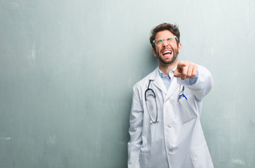 Young friendly doctor man against a grunge wall with a copy space shouting, laughing and making fun of another, concept of mockery and uncontrol