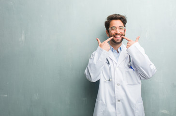 Young friendly doctor man against a grunge wall with a copy space smiles, pointing mouth, concept of perfect teeth, white teeth, has a cheerful and jovial attitude