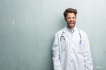 Young friendly doctor man against a grunge wall with a copy space screaming angry, expression of madness and mental instability, open mouth and half-opened eyes, madness concept