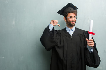 Young graduated man against a grunge wall with a copy space proud and confident, pointing fingers, example to follow, concept of satisfaction, arrogance and health