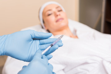 selective focus of syringe holding by beautician in front of woman face at beauty salon