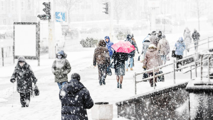 Blizzard in an Urban Environment. Silhouette of a Woman with a Red Umbrella in a Crowd of Rushing People in Snowfall. Abstract Blurry Winter Weather Background.