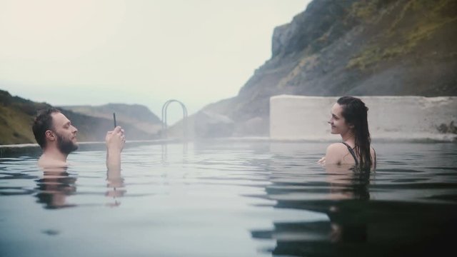 Young happy couple resting in hot springs in Iceland. Handsome man taking photos on smartphone of woman.