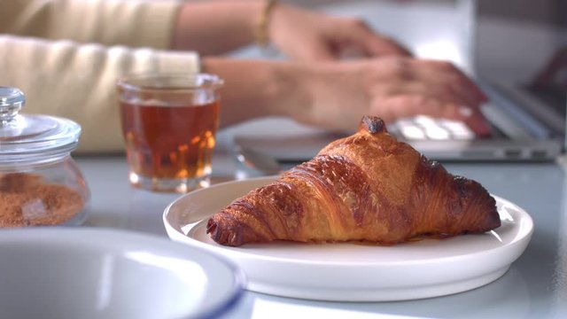womans hand working with notebook, focus on first plane with croissant and tea. Then womans hand take glass with tea