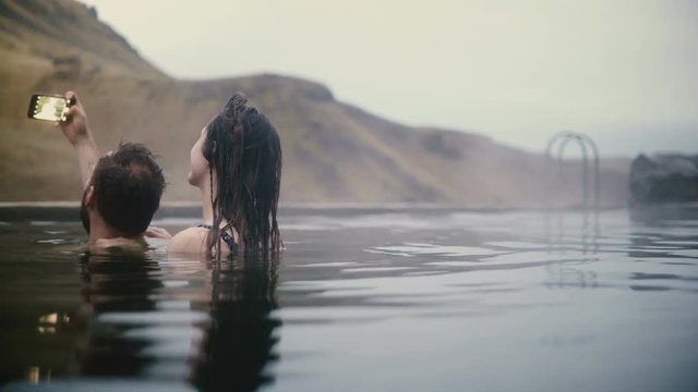 Young couple taking selfie photos on smartphone in mountains valley. Man and woman swimming in hot springs in Iceland.