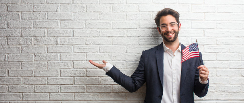 Young business man wearing a suit against a white bricks wall holding something with hands, showing a product, smiling and cheerful, offering an imaginary object - Powered by Adobe