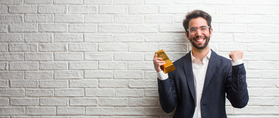 Young business man wearing a suit against a white bricks wall very happy and excited, raising arms,...