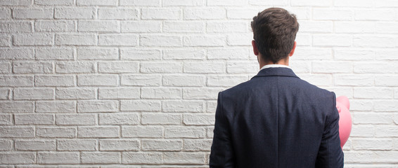 Young business man wearing a suit against a white bricks wall showing back, posing and waiting, looking back