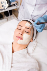 beautician applying cosmetic mask on woman face with cosmetic brush and holding vessel at beauty salon