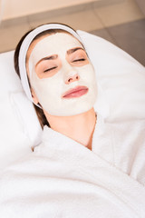 woman lying in white bathrobe with applied facial mask and closed eyes at beauty salon