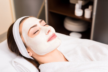 woman lying in white bathrobe with applied facial mask, smiling and closed eyes at beauty salon