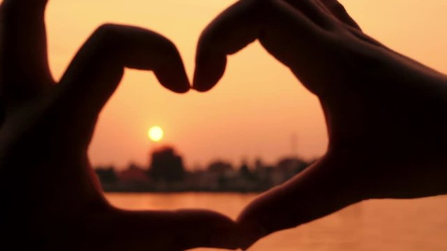 The shadow of a woman's hand made a heart shape and the sun is falling beside the shape and near the colorful river area.
