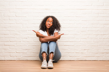 Young black woman sitting on a wooden floor confused and doubtful man, decide between two options, concept of indecision
