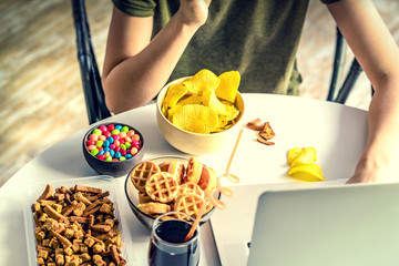 Girl works at a computer and eats fast food. Unhealthy food: chips, crackers, candy, waffles, cola. Junk food, concept.