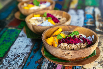 Close up smoothie bowl made with mango, banana, granola, grated coconut, dragon fruit, chia seeds and mint on colored background.
