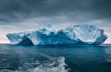 Fototapeta na wymiar Big massive iceberg floating among the frozen ocean. Antarctica epic scenery in blue and grey tints. The dark cloudy sky over the South pole. The wild harsh environment. Mysterious winter landscape.