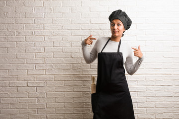 Young baker indian woman against a bricks wall proud and confident, pointing fingers, example to...