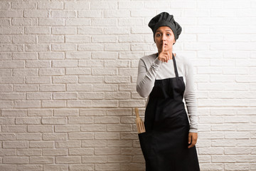 Young baker indian woman against a bricks wall keeping a secret or asking for silence, serious face, obedience concept