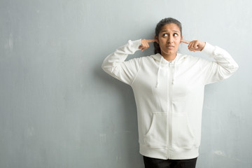 Young sporty indian woman against a gym wall covering ears with hands, angry and tired of hearing some sound