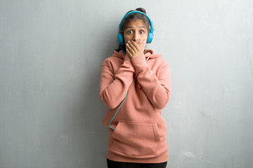 Fototapeta premium Young sporty indian woman against a wall covering mouth, symbol of silence and repression, trying not to say anything. Listening music with headphones.