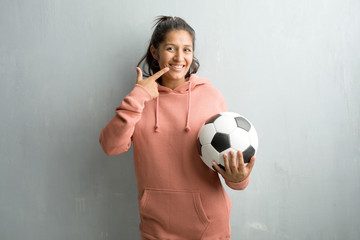 Young sporty indian woman against a wall smiles, pointing mouth, concept of perfect teeth, white teeth, has a cheerful and jovial attitude. Holding a soccer ball to play a game.